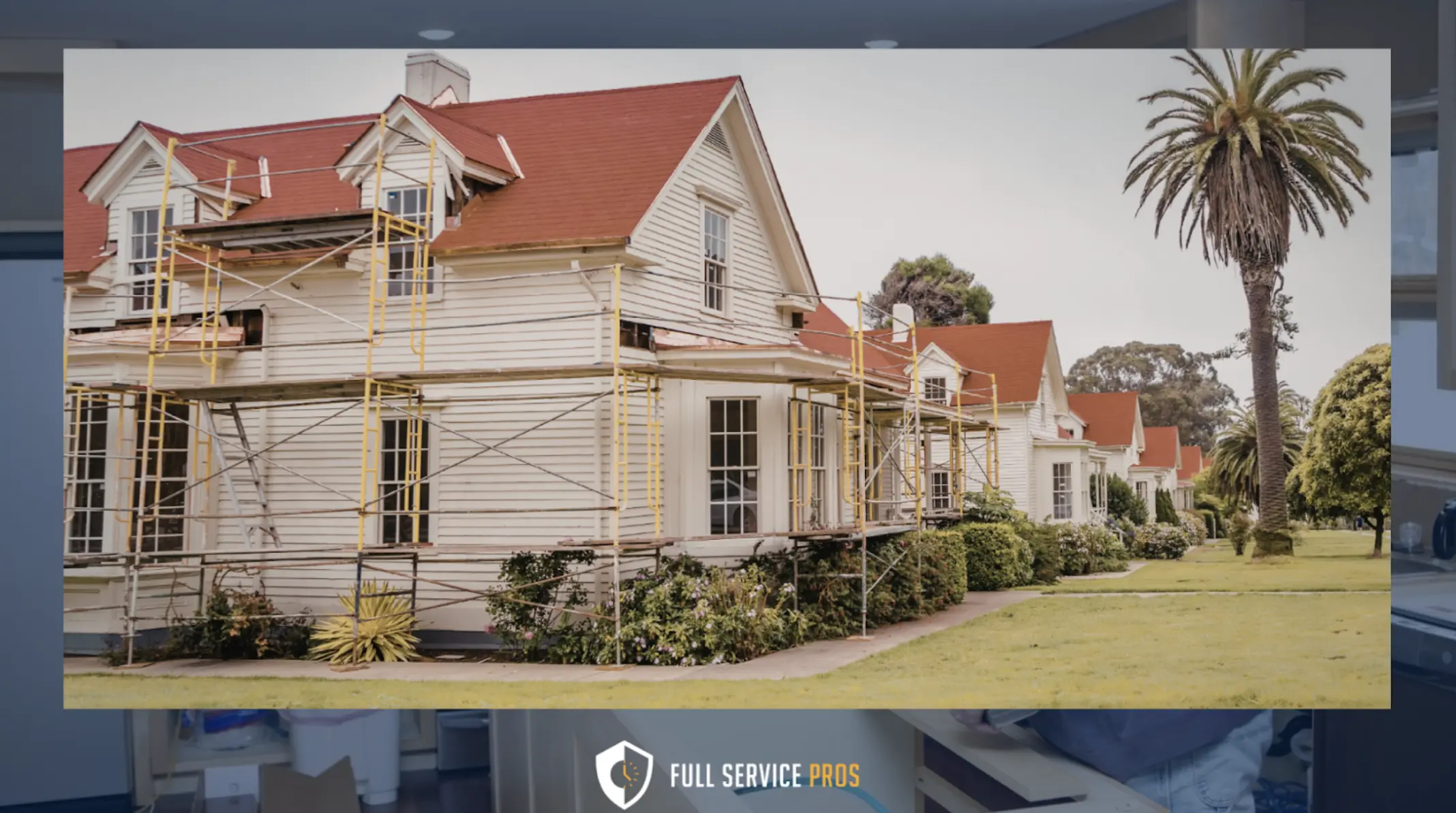 Restoration Services: A large white house with red roofs is surrounded by scaffolding for exterior repairs or painting; palm trees and grass are visible in the yard.
